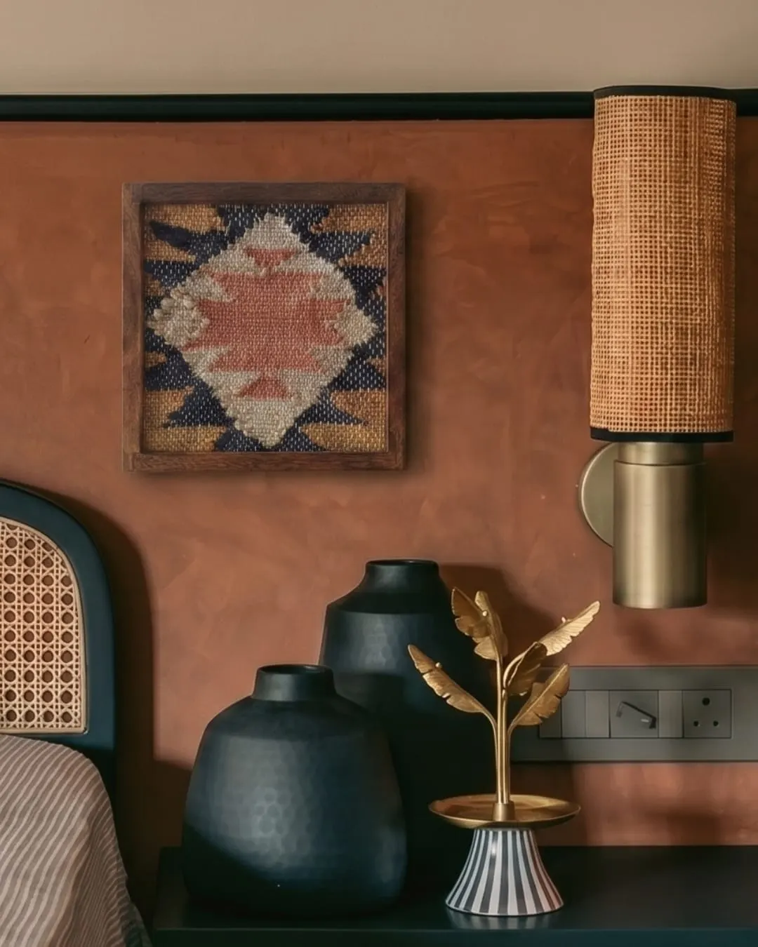 A framed hand-woven piece with a rust and navy diamond pattern on a terracotta wall above a bedside lamp
