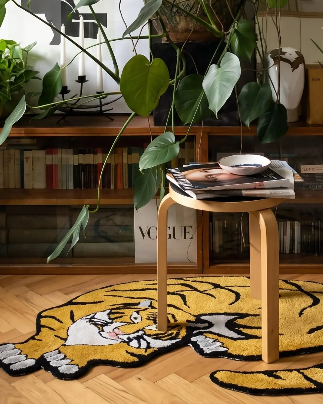 A tiger-shaped tufted rug on a herringbone floor beside a stool, with a bookcase behind