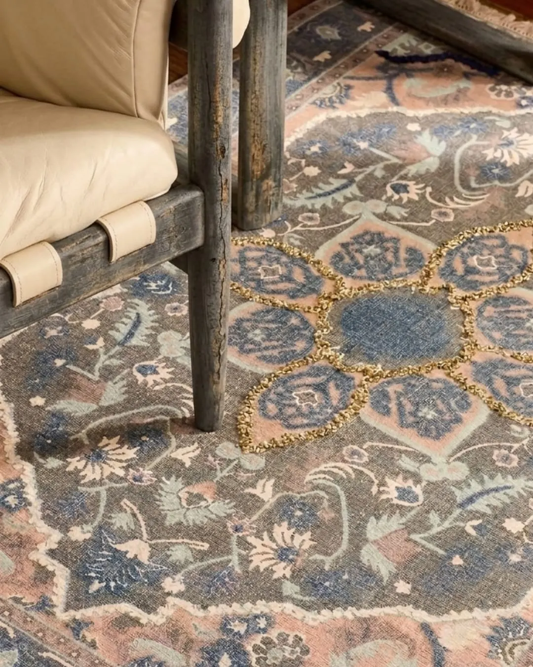 A muted floral rug in blue, rose and gold under a wooden chair with a cream cushion